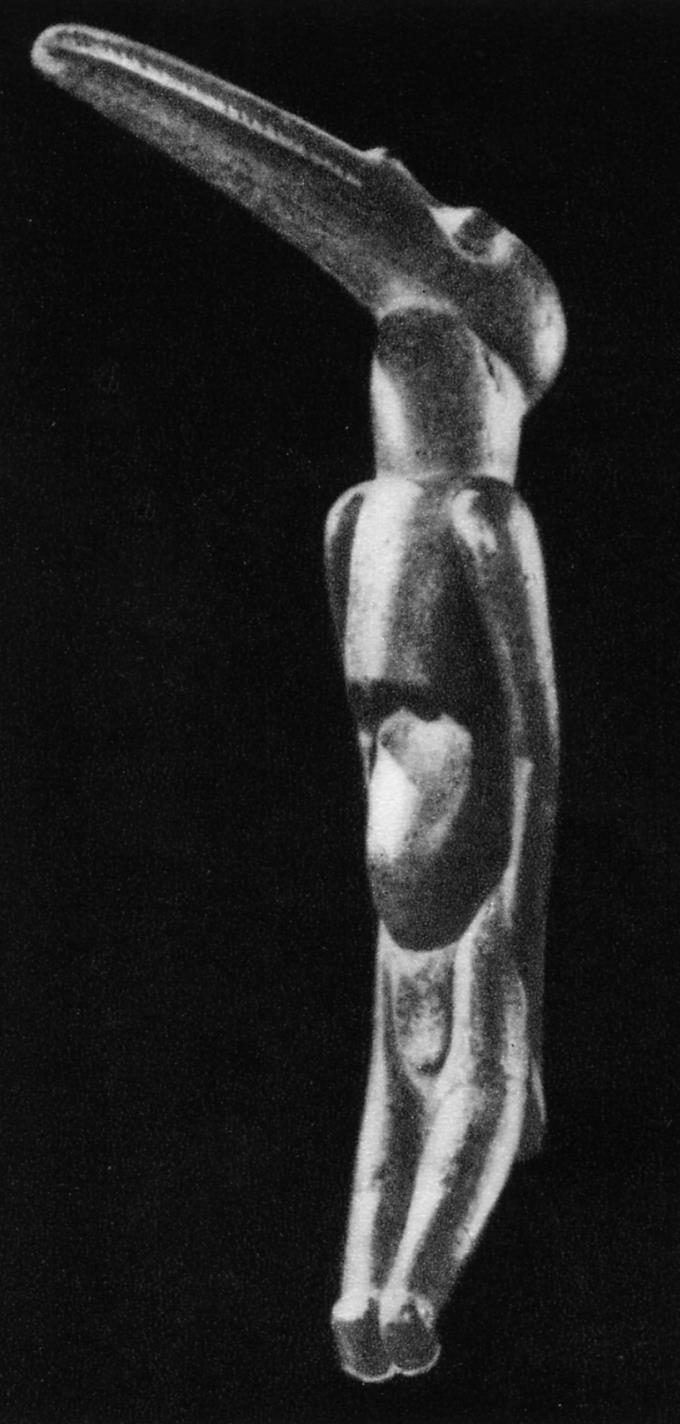 A carved wooden figure photographed upright against a black ground, lit from the left. It has a human body with long straight legs and arms held close to the sides, and in place of a human head a long curved beak sweeping up and back from a rounded skull. The polished wood catches the light along the beak, shoulders and shins. No surface markings are legible at this size.