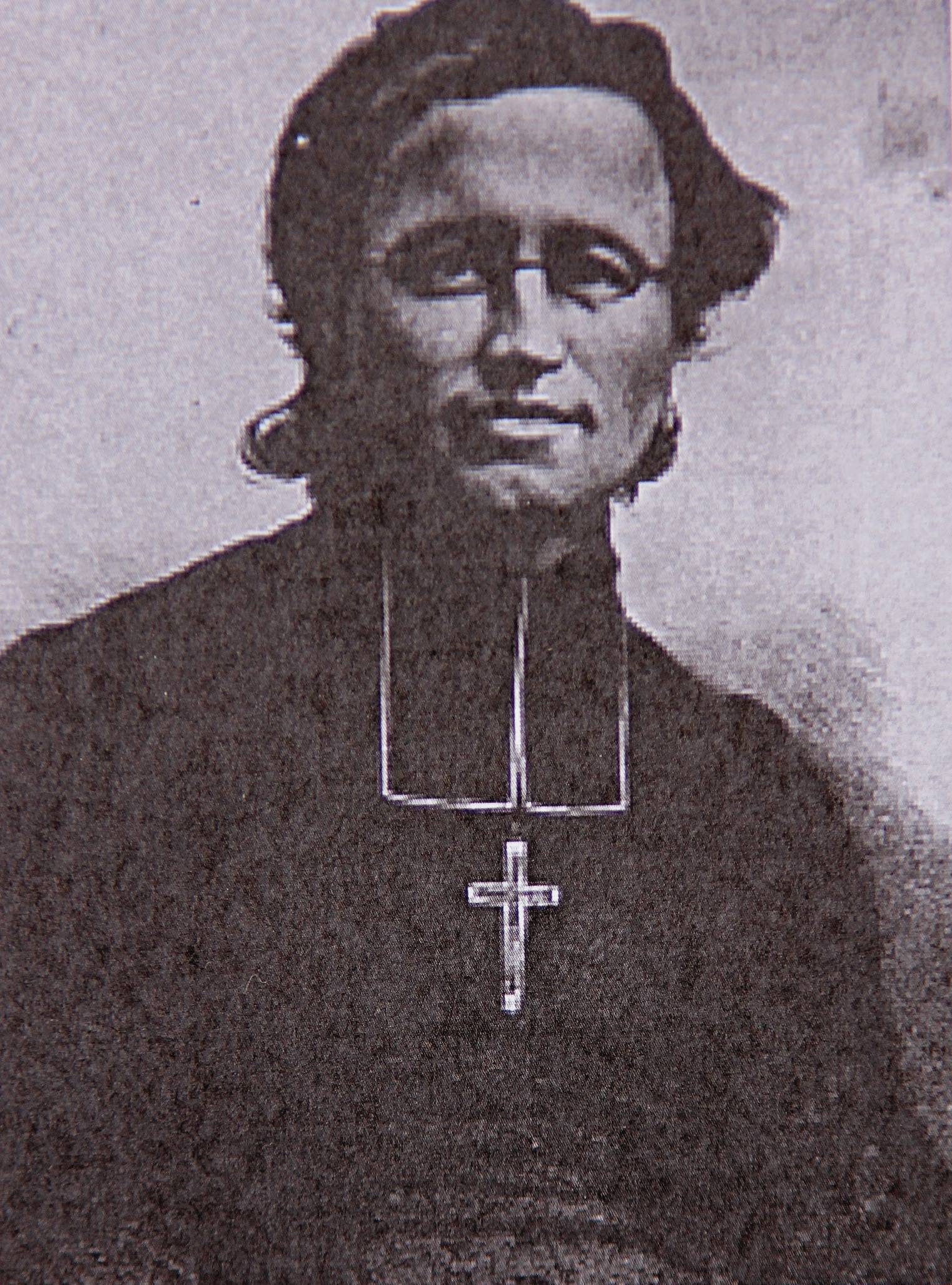 A head and shoulders portrait of an older man against a plain pale ground, in dark clerical dress with a pale bib at the throat and a plain pectoral cross hanging on a chain over his chest. His hair is swept back from a broad forehead and his mouth is set in a faint smile. The picture is a heavily grained monochrome reproduction, coarse enough that the texture breaks up across the face.