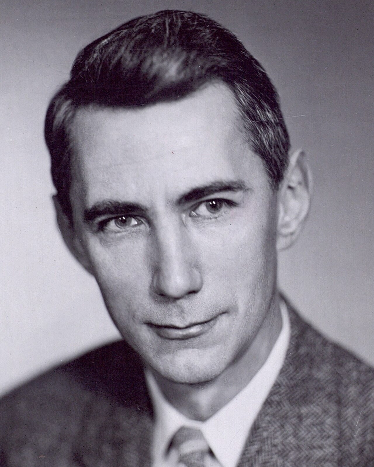 A black-and-white studio portrait photograph of Claude Shannon as a younger man, with dark hair combed back, wearing a tweed jacket and tie, against a plain background