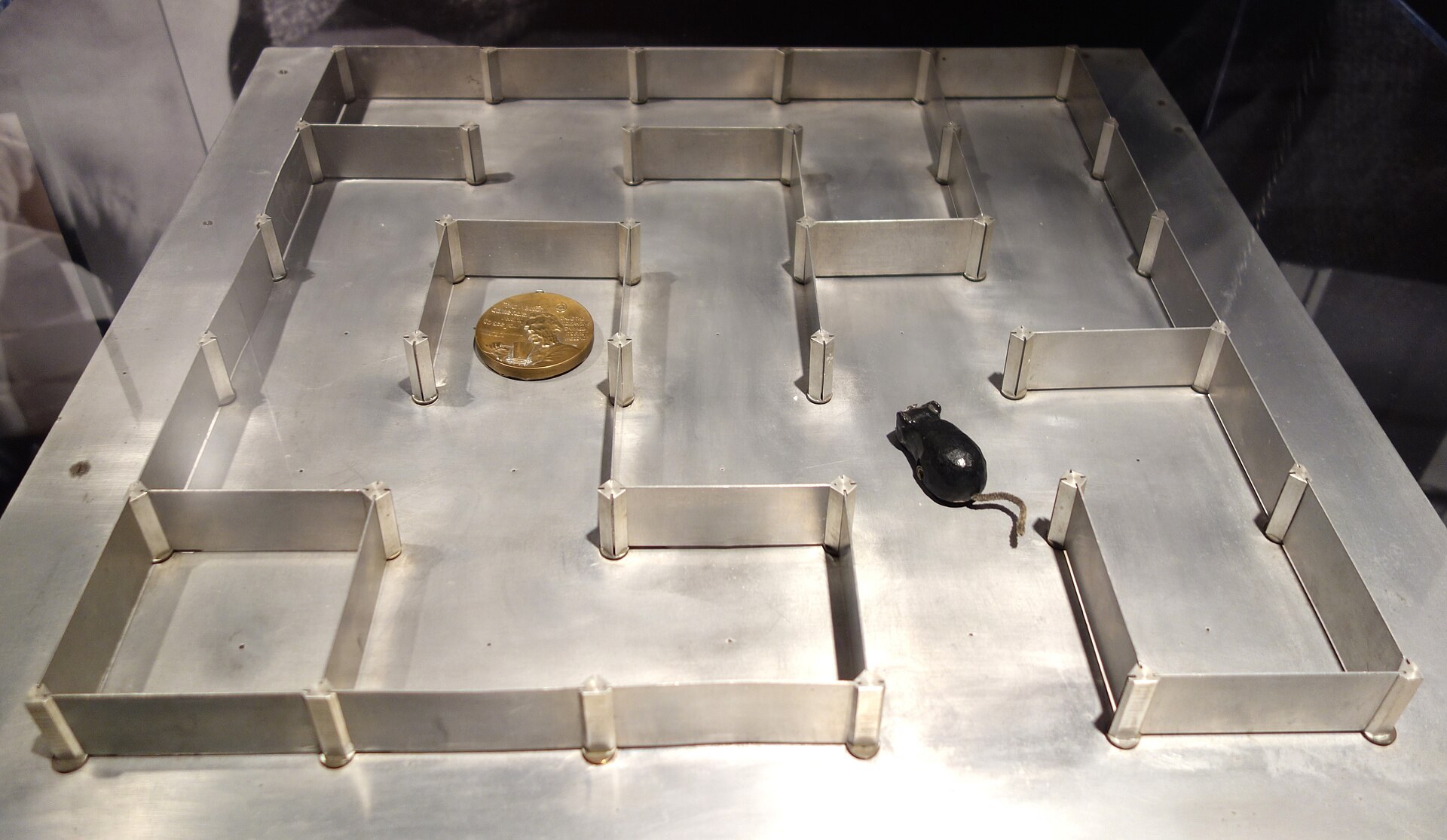 A photograph of a metal maze with movable partition walls on a metal base, a small black mechanical mouse with a string tail sitting in one of the passages, displayed at a museum