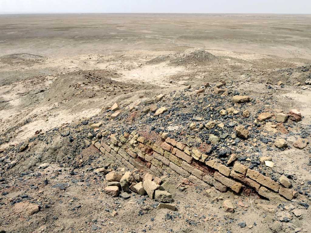 A low eroded mound under a pale hazy sky. Across the middle of the frame runs a single exposed course of baked brick, several bricks deep and clearly coursed, with broken brick and dark slag scattered over the surface around it. Beyond the mound the ground is flat empty desert to a hazy horizon. There is no structure, no excavation trench and nobody in the frame.