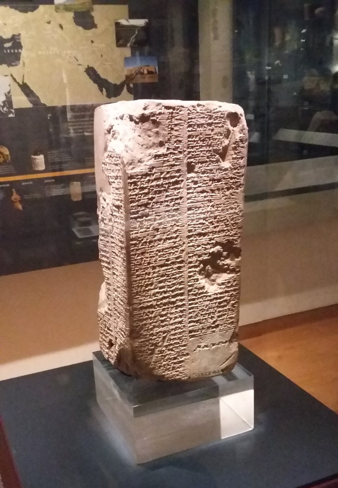A four-sided clay prism standing on a clear perspex block inside a glass museum case. Two columns of dense cuneiform cover the visible face, divided by a ruled vertical line. The upper left corner is chipped away and there is a large gouge at the upper right and another break at the lower right. Behind and reflected in the case glass, a gallery is visible with a wall map and a timeline.