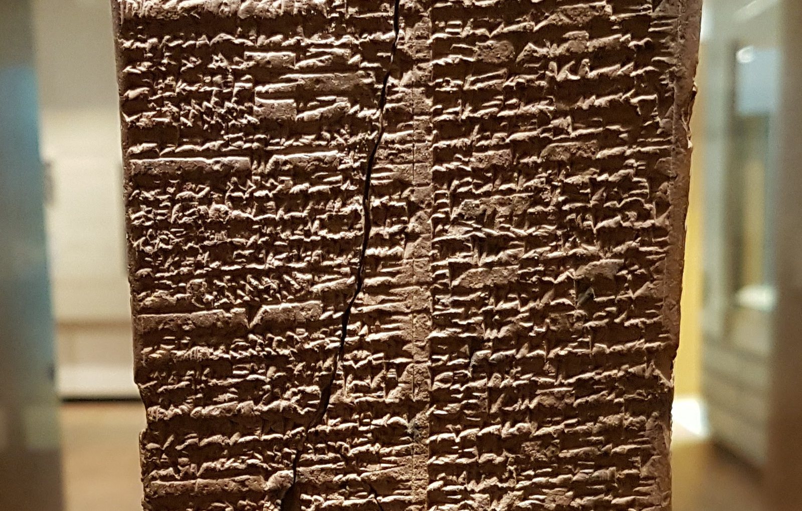 A close view of warm reddish-brown clay under raking light, so that each cuneiform wedge stands proud of the surface. Two columns of dense signs are separated by a clear blank vertical margin. Horizontal ruled lines divide the text into bands. A long jagged vertical crack runs the full height of the left column.