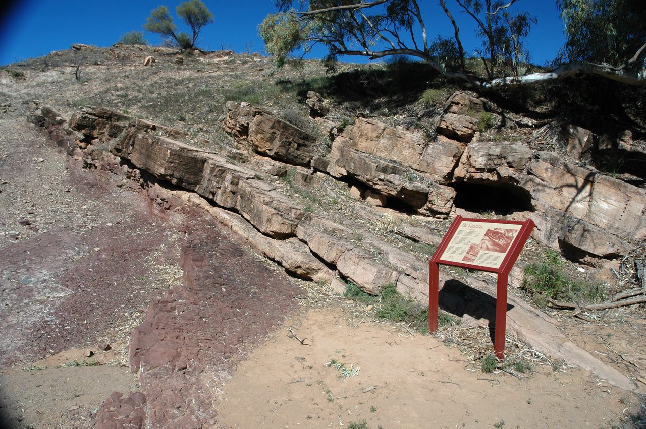A sunlit hillside outcrop under deep blue sky, a low ledge of pale pinkish tan rock in thin flat beds stepping across the slope, a broad apron of dull maroon red broken rock and rubble below it, and a small interpretive sign in a red steel frame standing on bare sandy ground