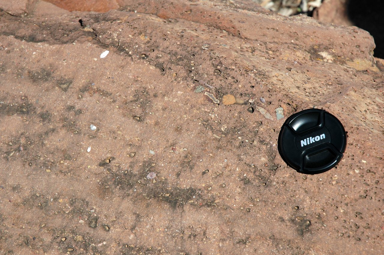 A broad weathered rock surface of dull reddish brown, densely peppered with small stones of many sizes and colours set at random in a fine matrix, with a black camera lens cap resting on it