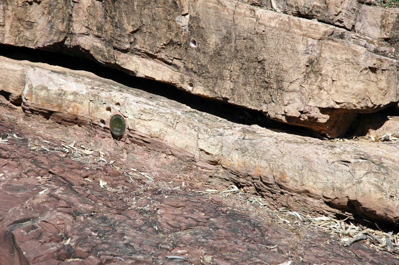 A close view of a sunlit rock face, pale bedded rock above and dull maroon red rock below, with several round cored holes bored into the pale beds and a dark olive green disc set flush into the face