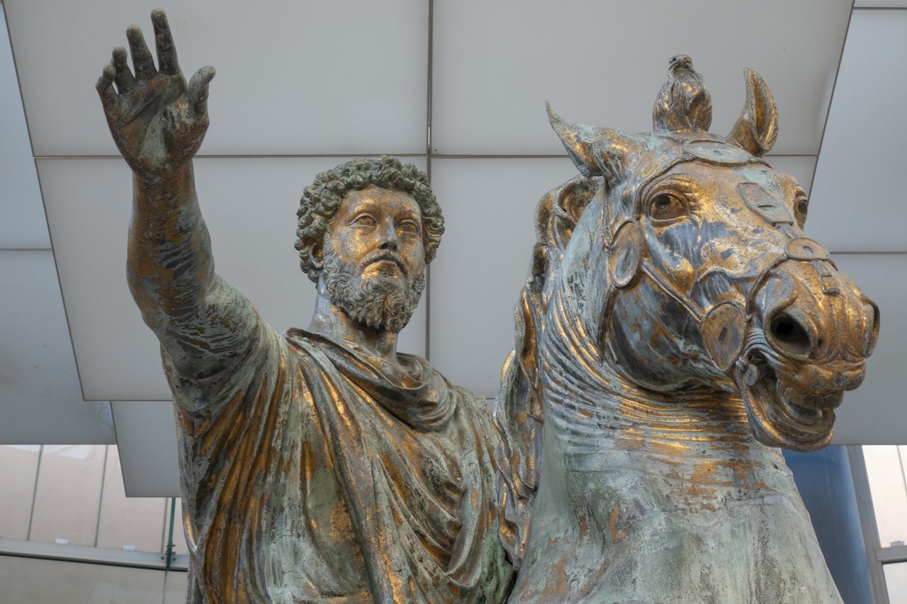 A close view from below of the original bronze equestrian statue of Marcus Aurelius, his right arm raised and the horse's head beside him, indoors against a modern white gallery ceiling