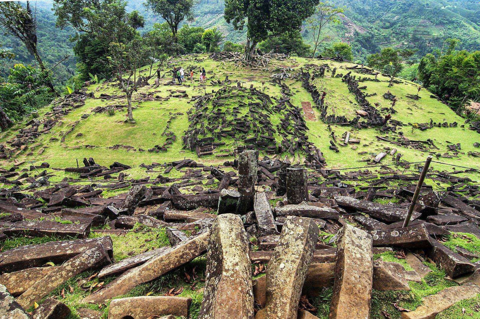 A hilltop terrace covered in thousands of loose stone columns. The blocks are long straight prisms with five and six sides, lying scattered and heaped in the foreground and arranged into low walls and rows on the grassy terraces above. A cluster stands upright near the centre foreground. Green mountains and forest fill the background, about six visitors stand on the upper terrace, and a wire fence and a wooden post cross the middle of the frame.