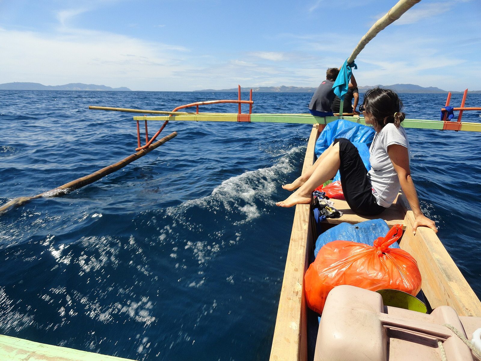 From inside a wooden outrigger boat under way in deep blue sea. Painted bamboo booms in green, red and yellow reach out to a bamboo float skimming the surface at the left, with a wake running back behind. Distant hills lie along the horizon. A woman in a white T-shirt sits on the gunwale at the right and two men are forward, with orange and blue plastic bags of luggage stowed in the hull.