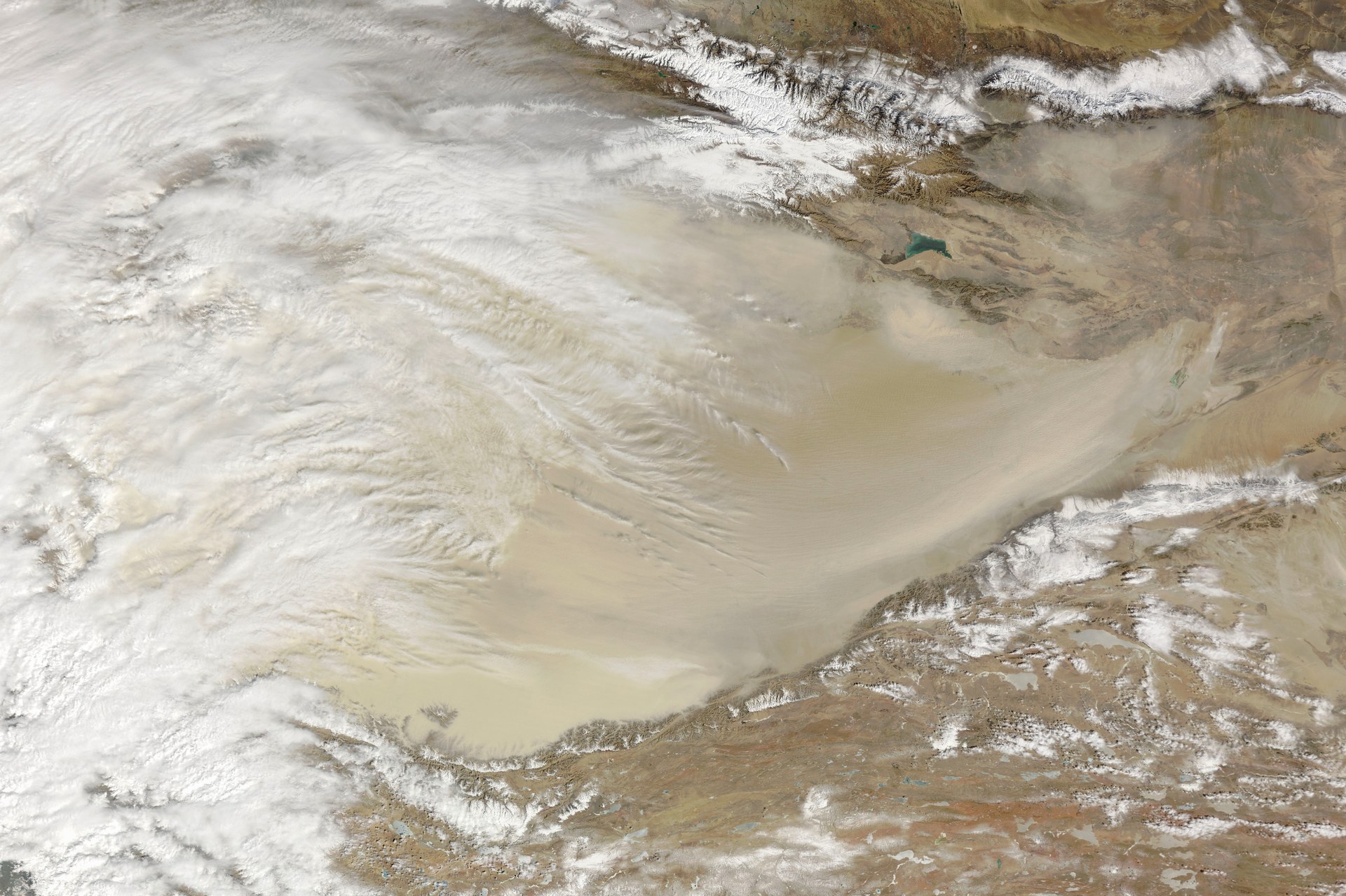 A satellite view of an elongated desert basin. A vast pale tan dust plume fills the centre and streams from left to right. Snow-covered mountain ranges run along the top of the frame and along the lower right, their white ridges sharp against brown highland terrain beyond. Thick white cloud spills in from the left. A small turquoise lake sits at the upper right.