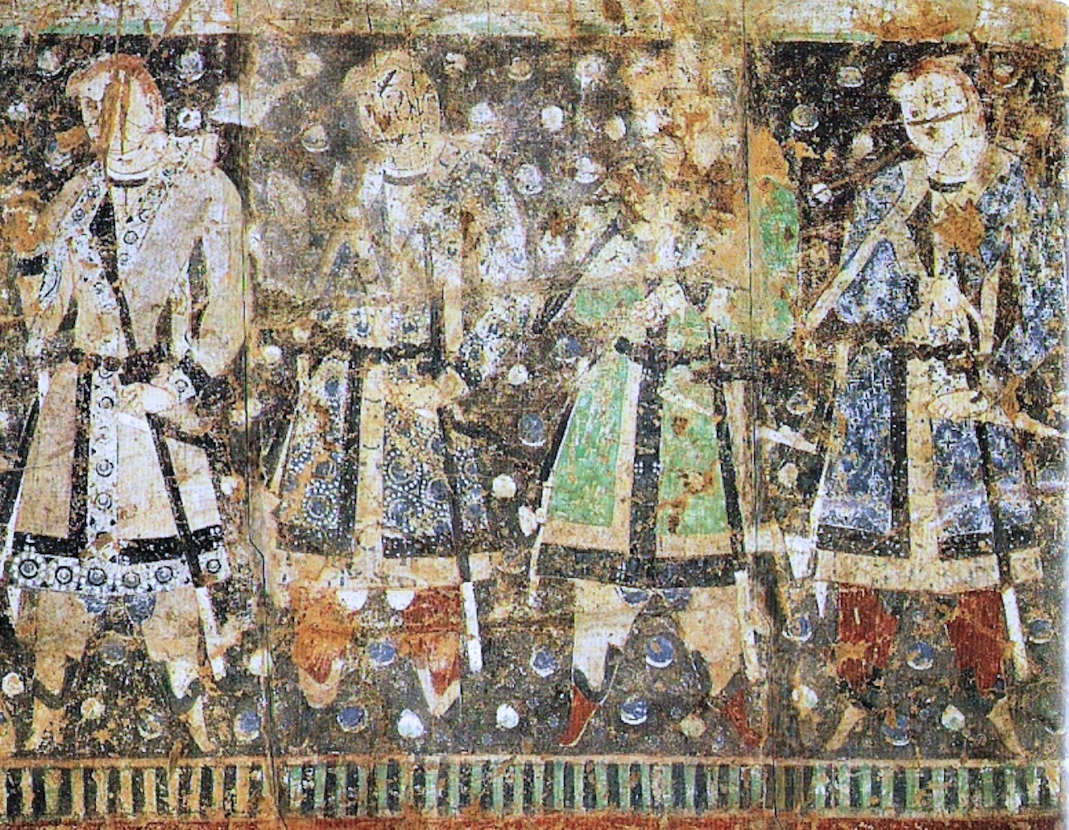 A weathered wall painting of four standing figures in a row, turned slightly to the left. They wear long belted knee-length coats with wide crossed lapels patterned with roundel medallions; one coat is green, the others cream and white with dark decorated panels. Broad belts carry hanging straps and what appear to be long swords. Red and orange leggings show below the coats above soft boots. The faces are blank pale ovals while the clothing detail around them remains crisp. The ground behind is dark and scattered with pale circular blobs, and a green and dark striped band runs along the bottom edge.