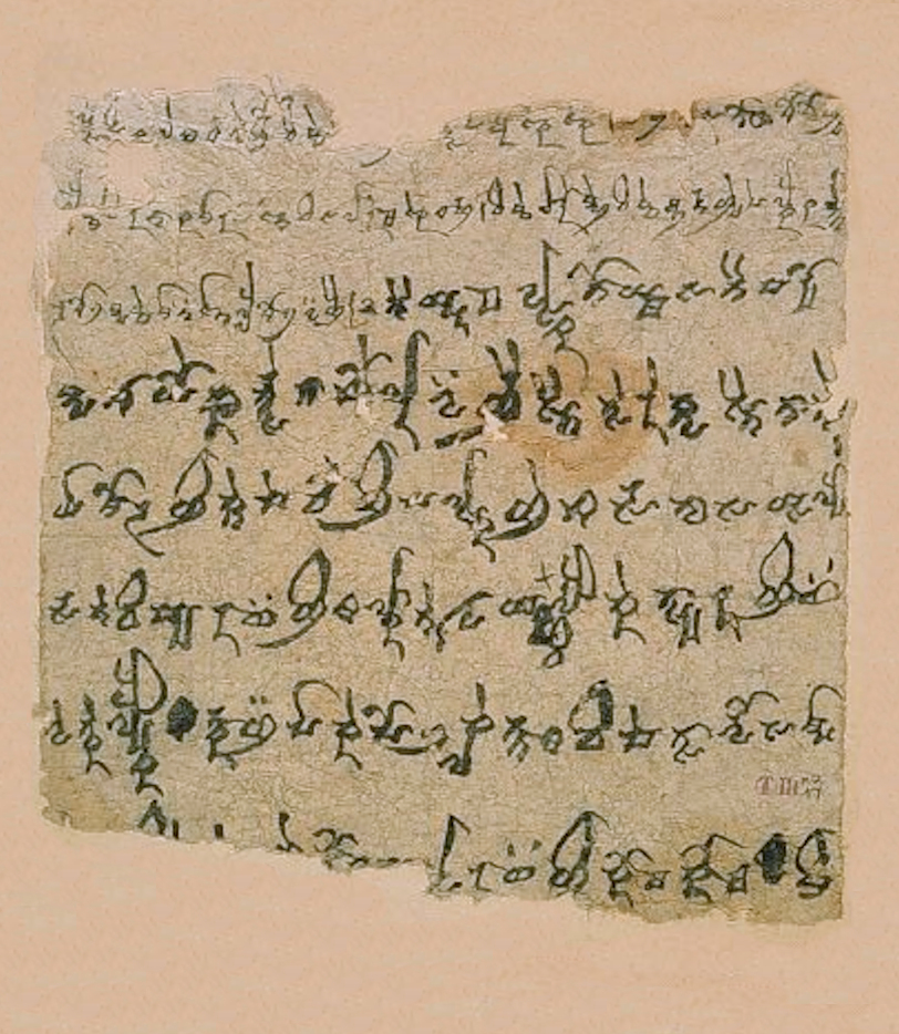 A ragged rectangular fragment of buff paper or parchment mounted on a pale pink backing board. About nine lines of dark brown-black ink in a flowing Brahmi-derived script run across it from left to right, with hooked and looped verticals. The top corners are torn away and a brownish stain sits in the upper middle right. A small faint catalogue mark appears at the lower right.