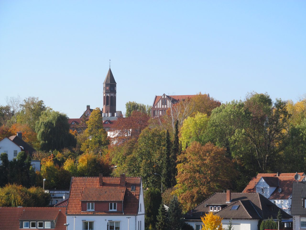 A colour photograph across the rooftops of Treysa in autumn: modern houses with red pantiled roofs in the foreground, a mass of turning trees behind them, and a red brick tower with a steep dark spire standing above the treeline under a clear sky
