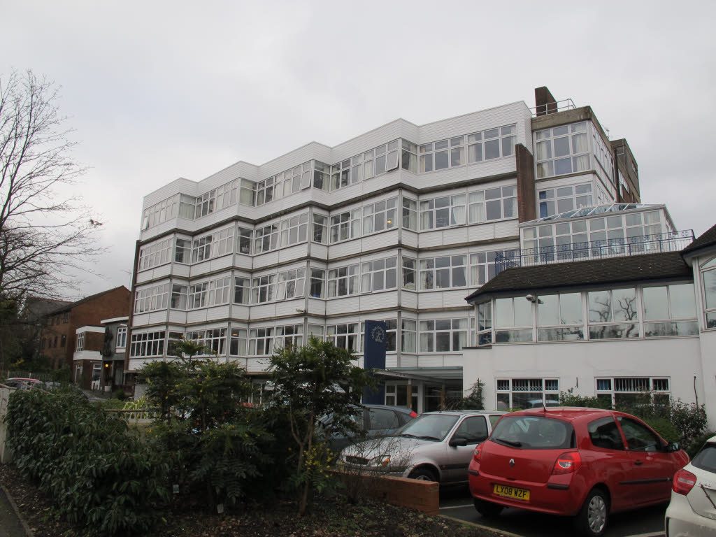 A colour photograph of St Christopher's hospice in Sydenham: a four-storey white building with near-continuous bands of windows around a faceted corner, a canopied entrance, and cars parked on the forecourt under an overcast sky