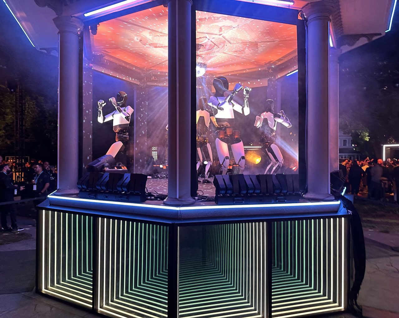 Humanoid robots inside a lit, columned pavilion at an evening promotional event, with a crowd standing outside it