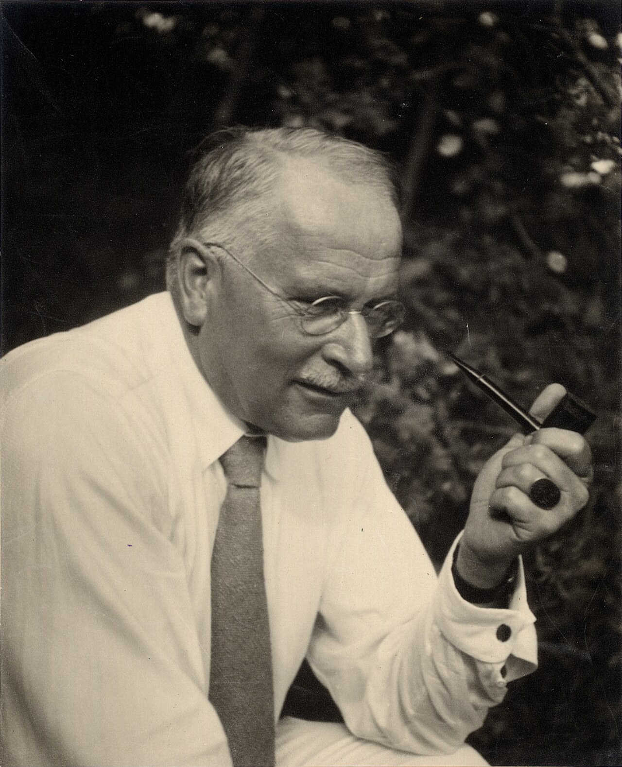 A sepia photograph of an elderly Carl Jung seated outdoors before dark foliage, wearing a light jacket and tie and round wire-rimmed spectacles, holding a smoking pipe up to the side of his face, a signet ring on his hand