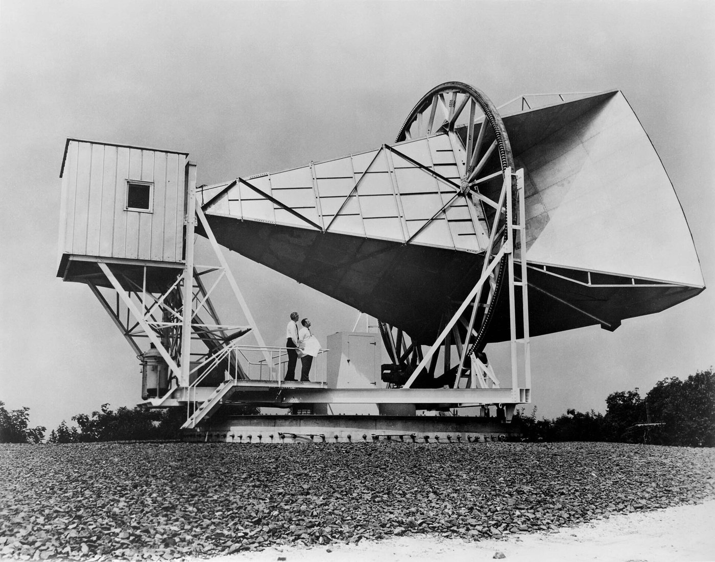 A black-and-white photograph of a large horn-shaped radio antenna, a metal scoop the size of a small building on a rotating mount, with two people standing on its platform for scale in a gravel field