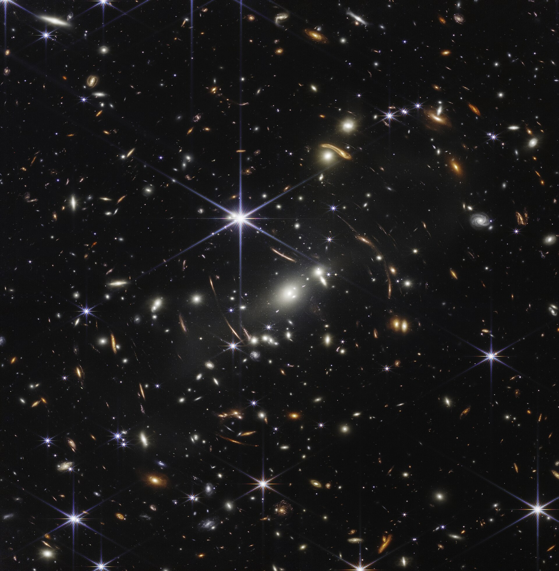 A James Webb Space Telescope deep-field image: a black field densely packed with thousands of galaxies of every shape and color, several bright foreground stars showing eight-pointed diffraction spikes, and curved, stretched arcs of light around a central cluster
