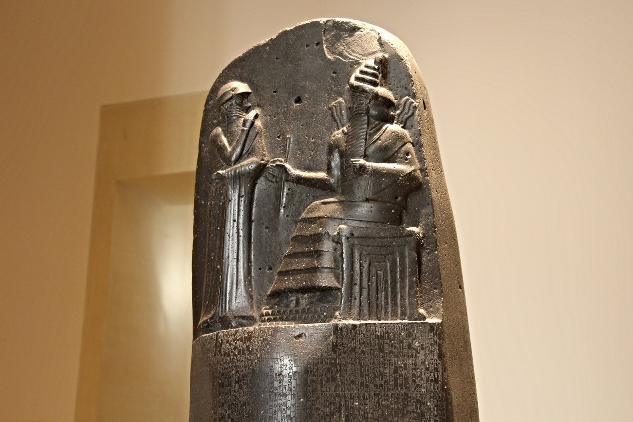 The top of the black basalt stele of the Code of Hammurabi, photographed in a museum: a carved relief of King Hammurabi standing with his hand raised before the enthroned sun-god Shamash, who wears a horned crown and radiates flames from his shoulders, with rows of cuneiform law beginning in the stone below