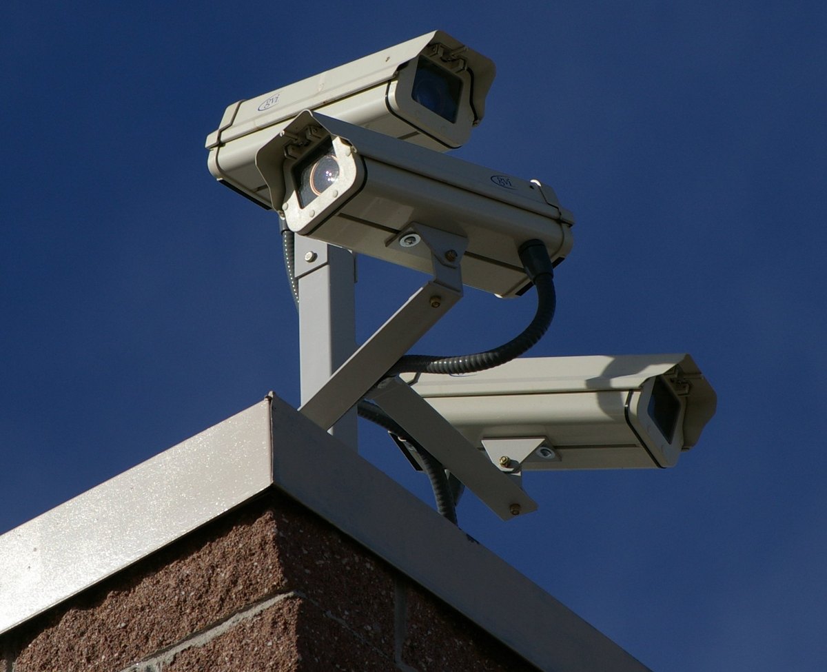 Three white CCTV surveillance cameras mounted together on a bracket at the corner of a brick building, each pointing in a different direction against a deep blue sky