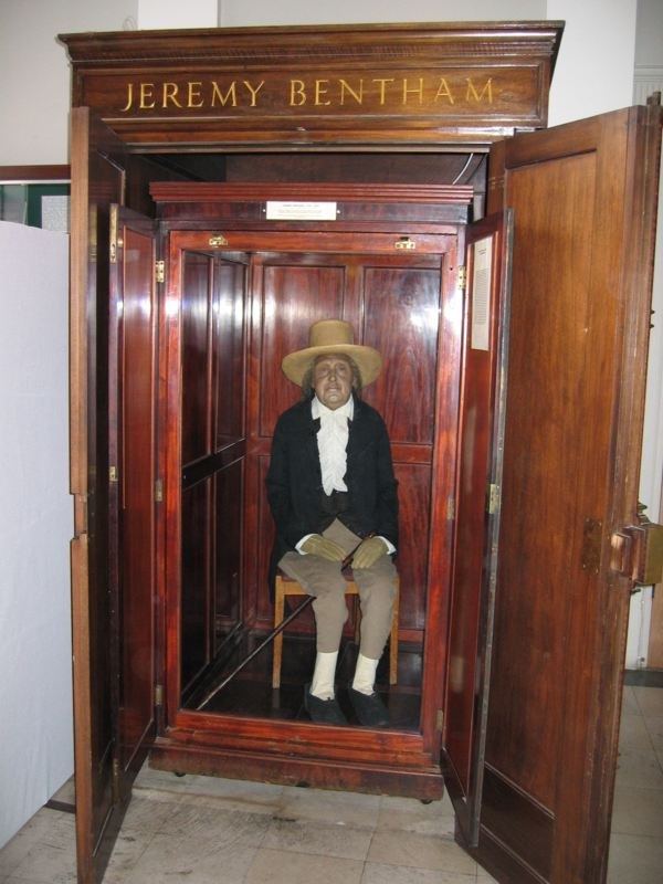 Jeremy Bentham's 'auto-icon': his preserved skeleton, dressed in his own dark coat and a wide-brimmed hat with a wax head, seated holding a walking stick inside an open wooden display cabinet labelled 'JEREMY BENTHAM'