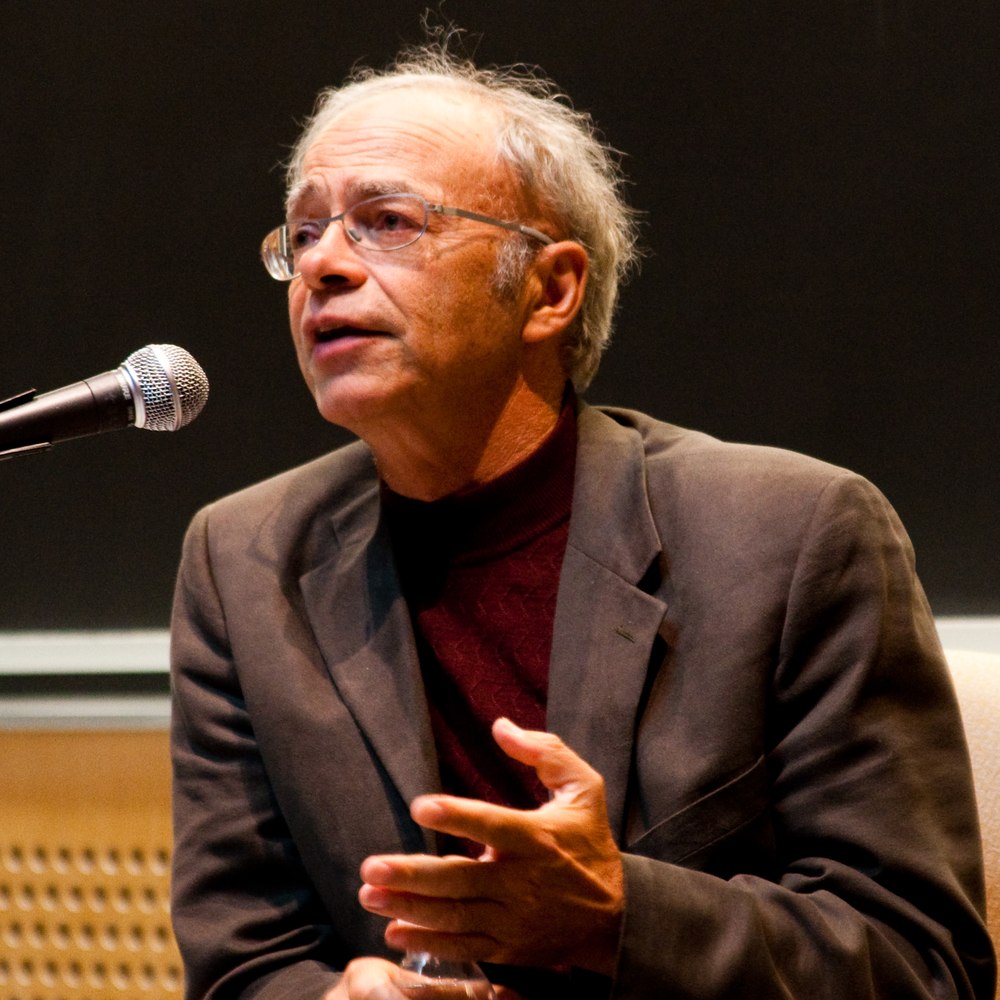 A photograph of the philosopher Peter Singer, grey-haired and wearing glasses and a brown jacket over a dark red turtleneck, speaking and gesturing at a microphone