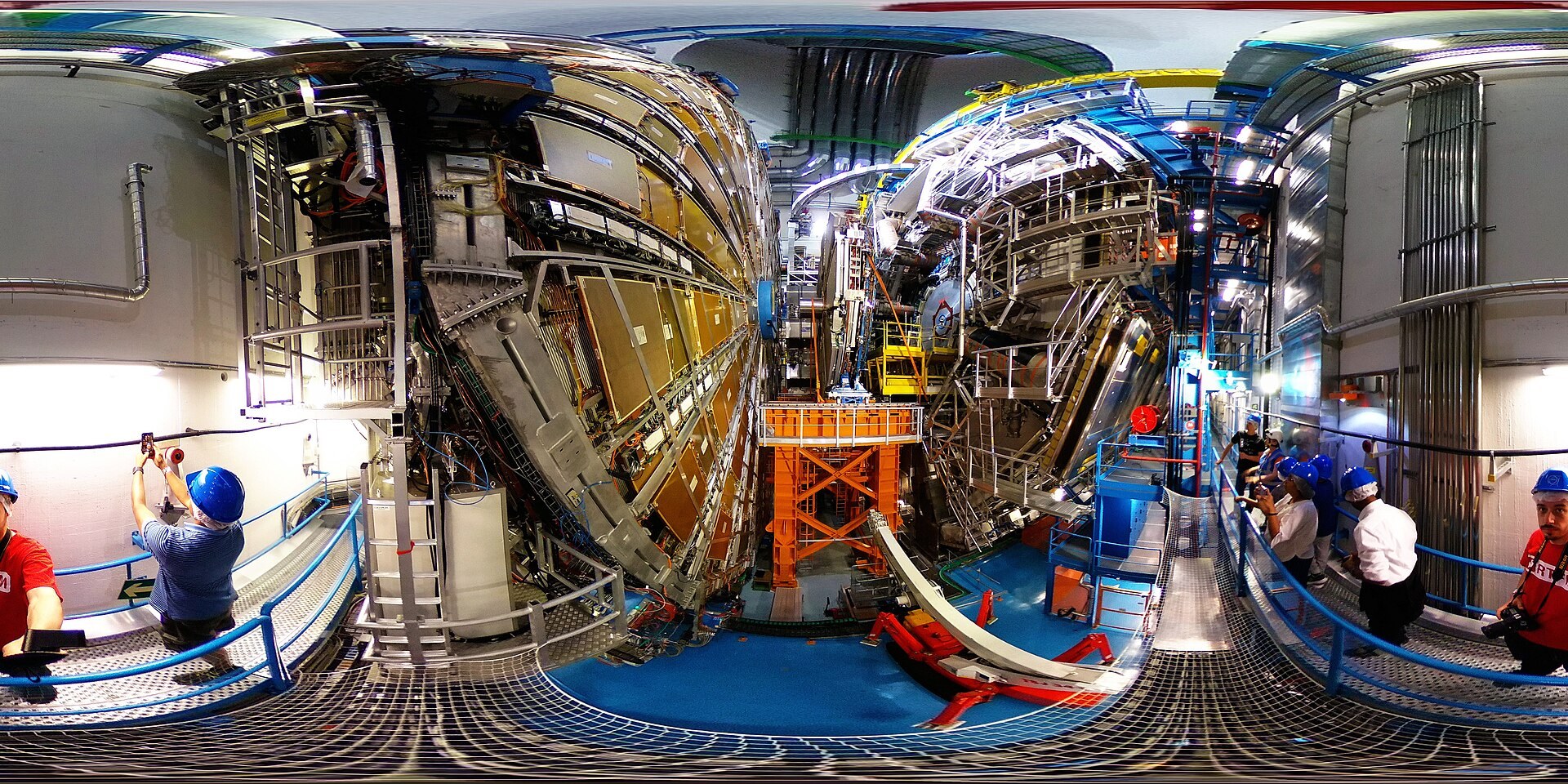 A wide interior photograph of the ATLAS particle detector at CERN: a vast, many-layered cylindrical machine of metal, cabling, and orange support structures filling a cavern, with people in hard hats on walkways at its base for scale