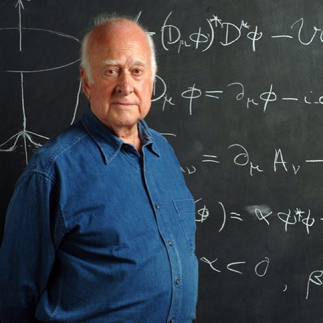 A photograph of an elderly Peter Higgs with white hair, in a blue denim shirt, standing in front of a blackboard covered with the chalk equations of the Higgs field and a small sketch of the Mexican-hat potential
