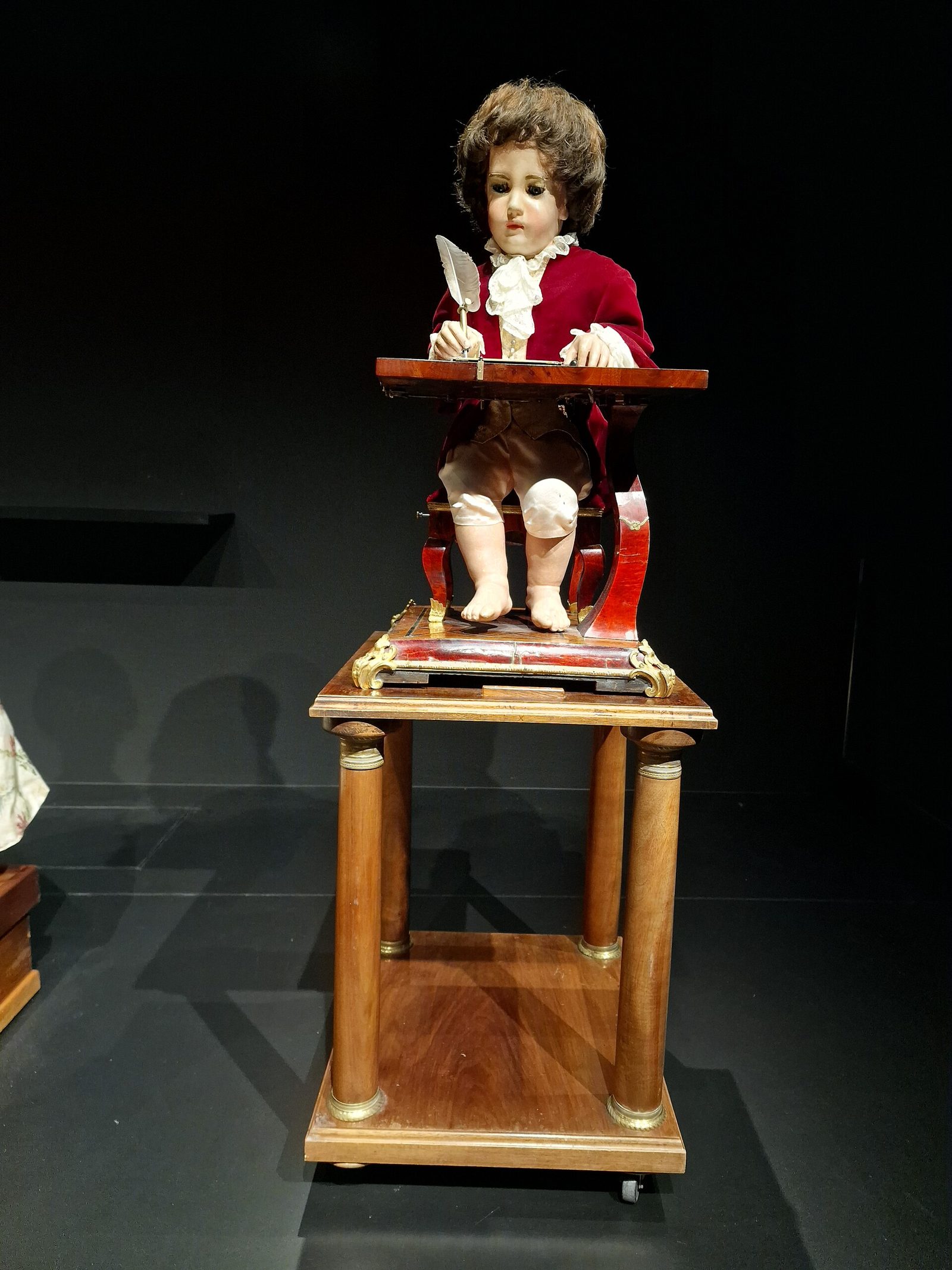 A small seated figure of a child in a red velvet coat with lace cuffs and a lace cravat, holding a quill pen over a writing surface, mounted on an ornate wooden plinth with turned columns and gilded fittings, displayed against a black museum ground. The figure's head is tilted toward the page and its free hand rests on the desk. The mechanism is enclosed in the base beneath it.