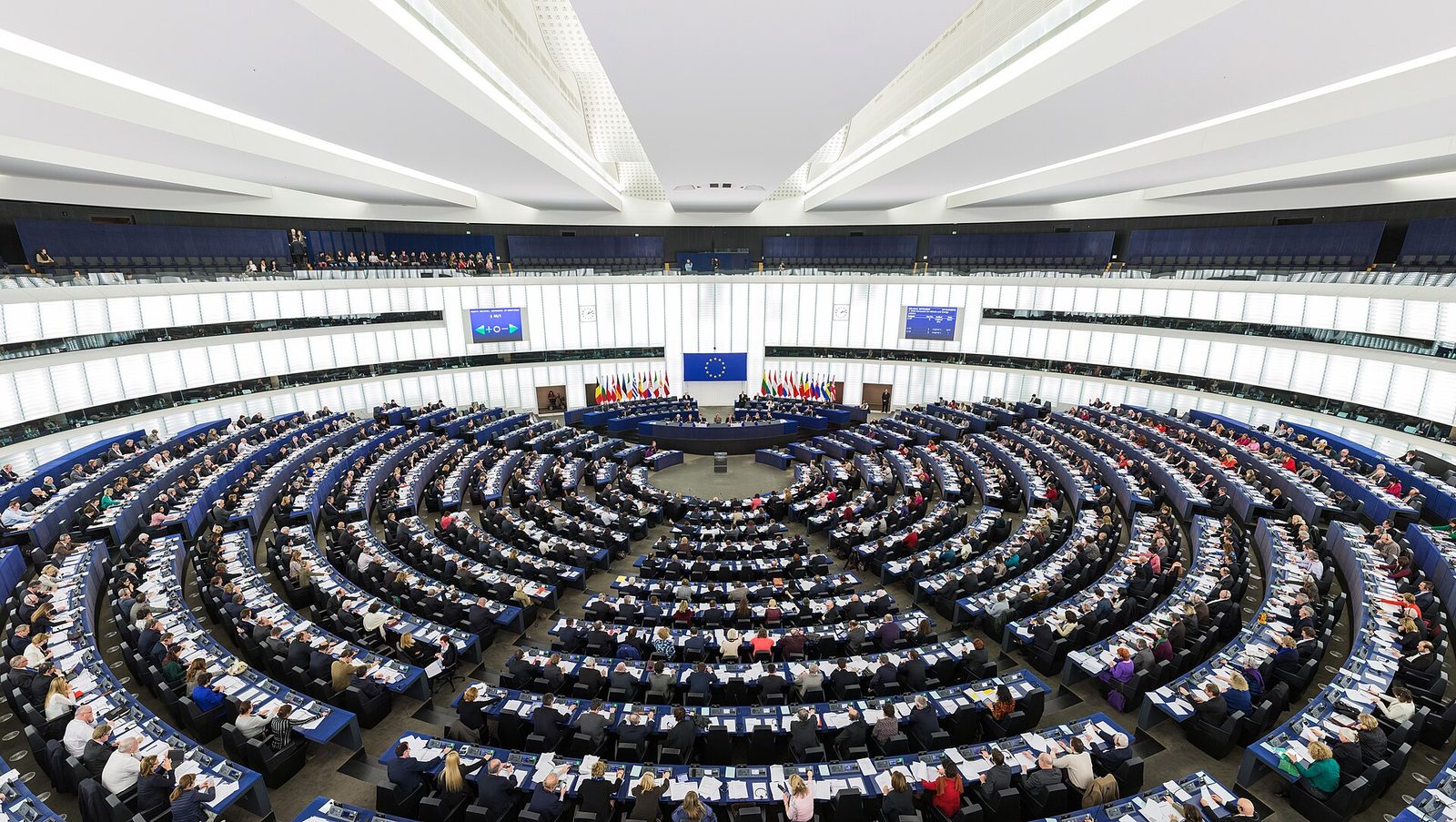 A wide interior view of a large circular debating chamber seen from above and behind. Concentric curved rows of blue desks fill the floor, every place occupied, sloping down toward a central podium. A row of national flags and a European Union flag stand along the far wall beneath two illuminated display screens showing a vote tally. Tiered public galleries run around the upper level, and long panels of white light form the ceiling.