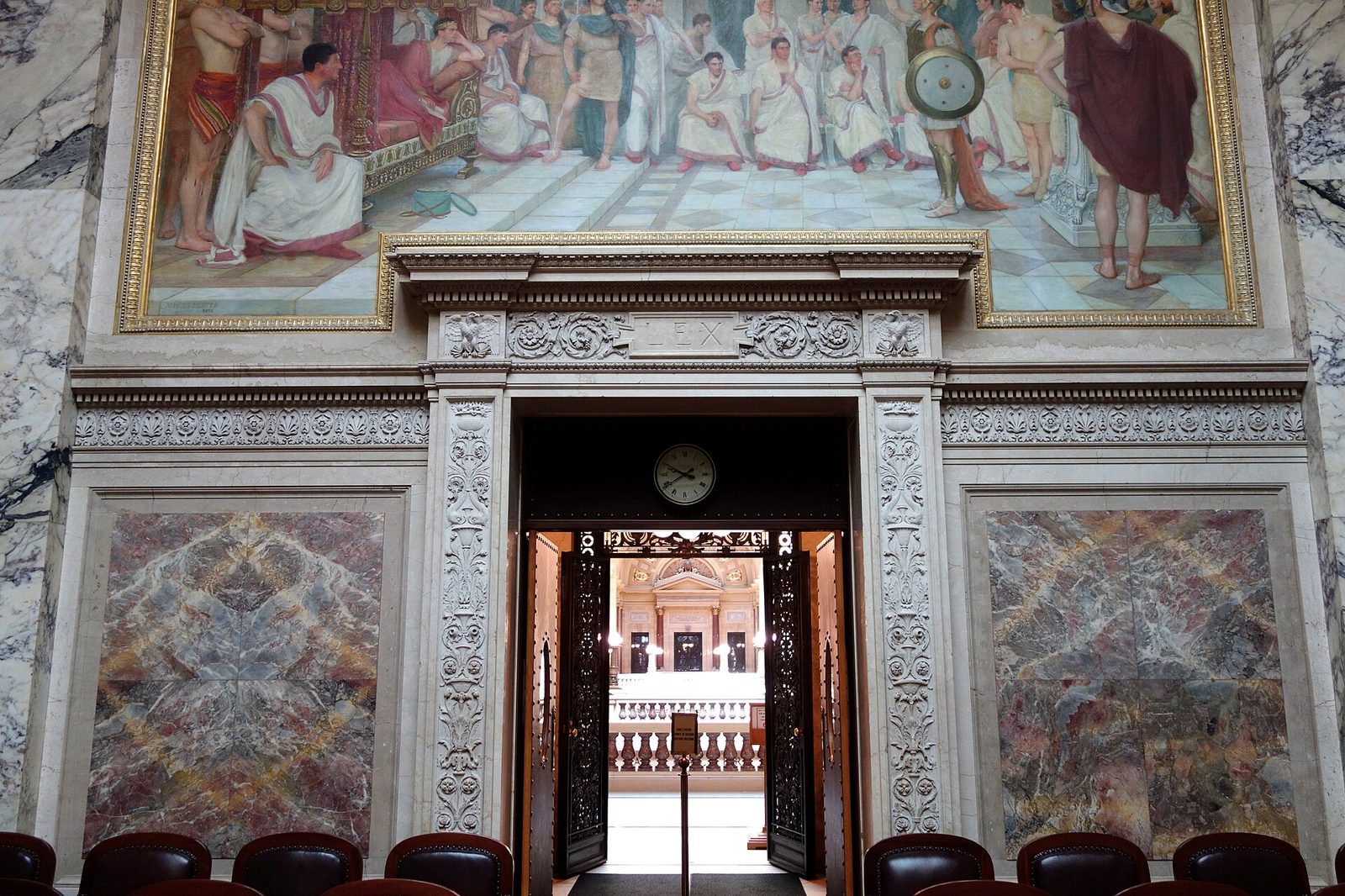 The interior of a marble courtroom photographed toward a tall carved stone doorway, its lintel inscribed with the word LEX between two eagles. Above the doorway hangs a large classical mural of robed figures in a colonnaded hall. Panels of veined marble flank the door, ornate metal doors stand open onto a lit space beyond with a round clock and a balustrade, and the tops of leather chairs cross the foreground. The room is empty of people.