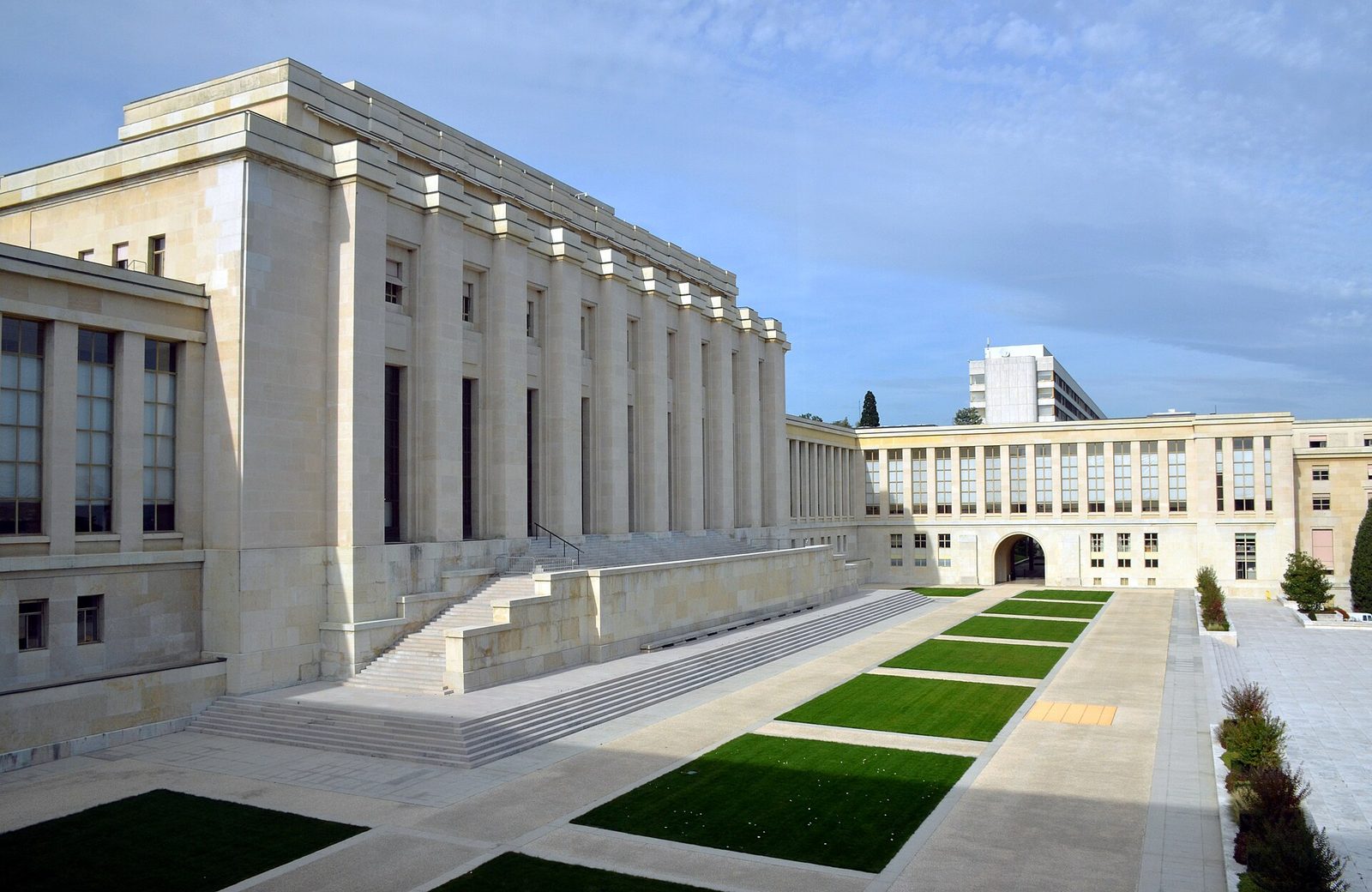 A long neoclassical stone building with a tall colonnade running along its front, photographed from an elevated angle in bright daylight under a partly clouded sky. A broad flight of steps descends from the colonnade to a paved courtyard laid with rectangular lawns. A lower wing with a long row of windows extends across the right of the frame. No people are visible.