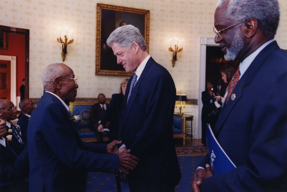 A color photograph of President Bill Clinton at the White House in 1997, clasping the hand of an elderly African American survivor of the Tuskegee Syphilis Study