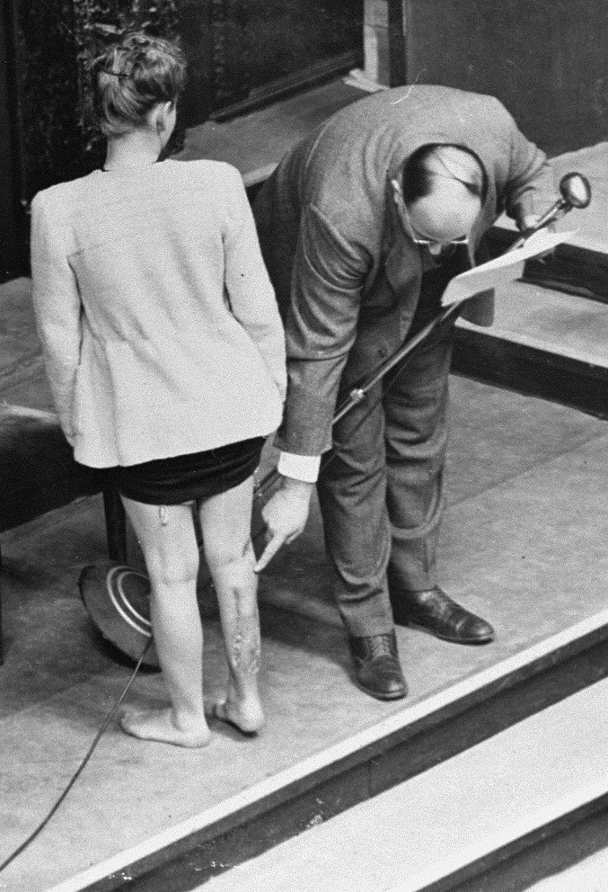 A black-and-white courtroom photograph seen from behind: Jadwiga Dzido, a young woman, stands with one lower leg bared to the tribunal while the physician Leo Alexander bends to point out the surgical scars left by the medical experiments performed on her