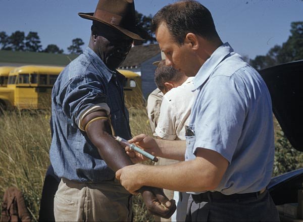 A color documentary photograph from the Tuskegee Syphilis Study: a Public Health Service worker drawing blood from the arm of an African American man, a participant, who stands outdoors in a field with a school bus behind them
