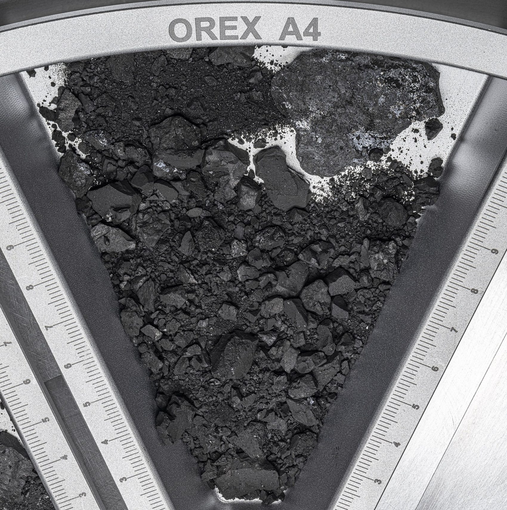 An overhead photograph of black, coal-like asteroid rubble and fine dark dust filling a wedge-shaped metal tray engraved with the label OREX A4, with ruler scale markings along the tray's straight edges