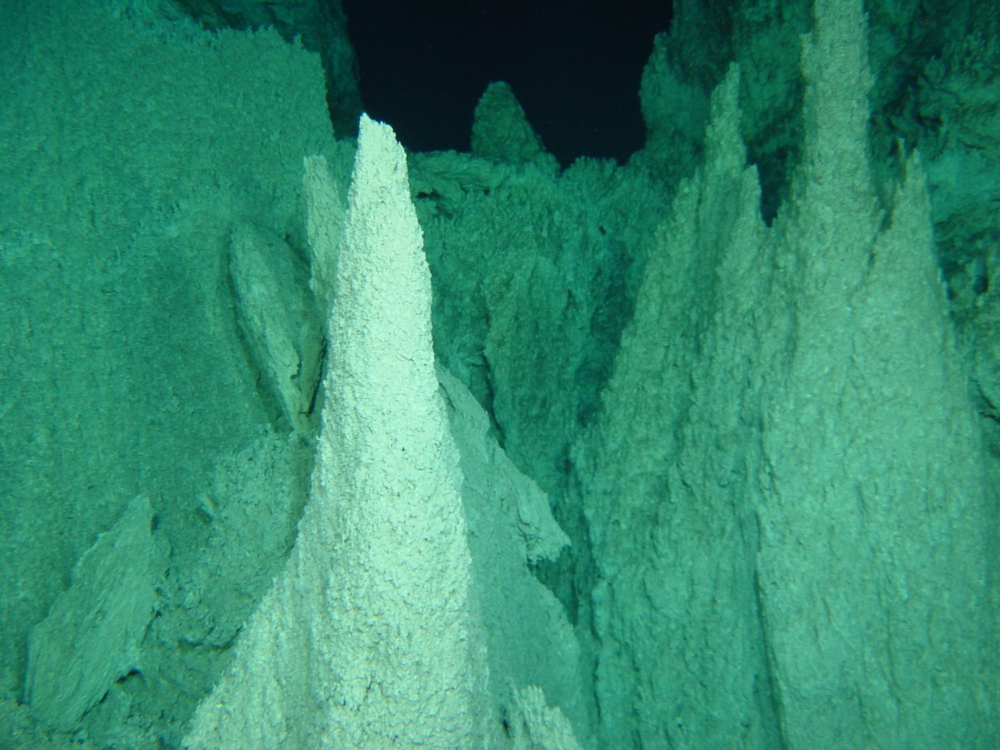 A deep-sea photograph of tall, pale, tapering carbonate mineral towers rising like spires against black water, lit greenish by an underwater vehicle's lights
