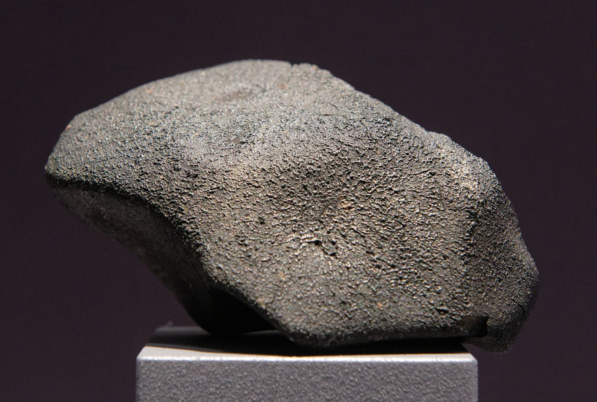 A dark grey, rounded, heavily pitted meteorite fragment resting on a pale grey museum pedestal against a deep maroon background, its crusty surface textured with fine bumps and hollows