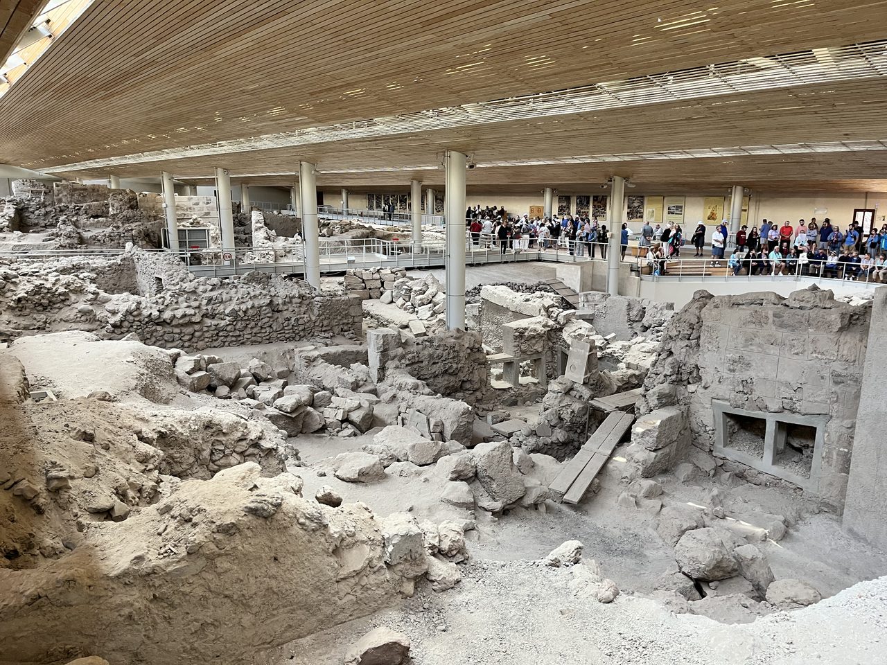 The excavated Bronze Age town of Akrotiri seen from above: pale stone walls, doorways and a preserved window opening under a large modern timber roof, with visitors and railings above