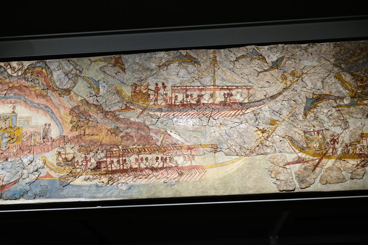 A long fresco panel behind glass showing oared ships with cabins and crews moving away from a single coastal town at the left edge, dolphins leaping around them, the painted surface visibly broken into fragments set in plain fill