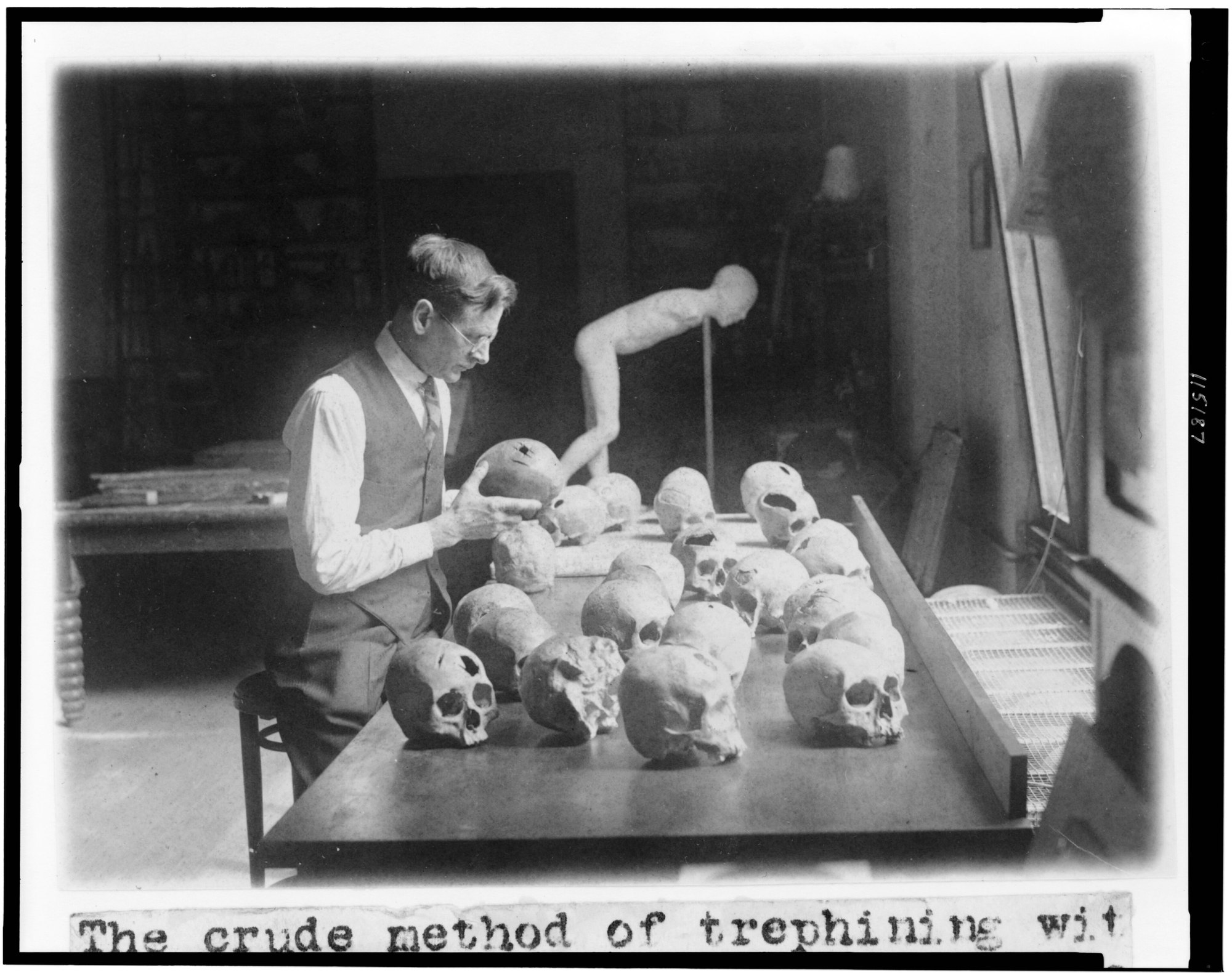 A black-and-white 1926 photograph of a man in a waistcoat and glasses seated at a table crowded with roughly a dozen or more human skulls, holding one up to examine an opening in it; museum shelves are visible behind, and a typed caption strip below reads 'The crude method of trephining'