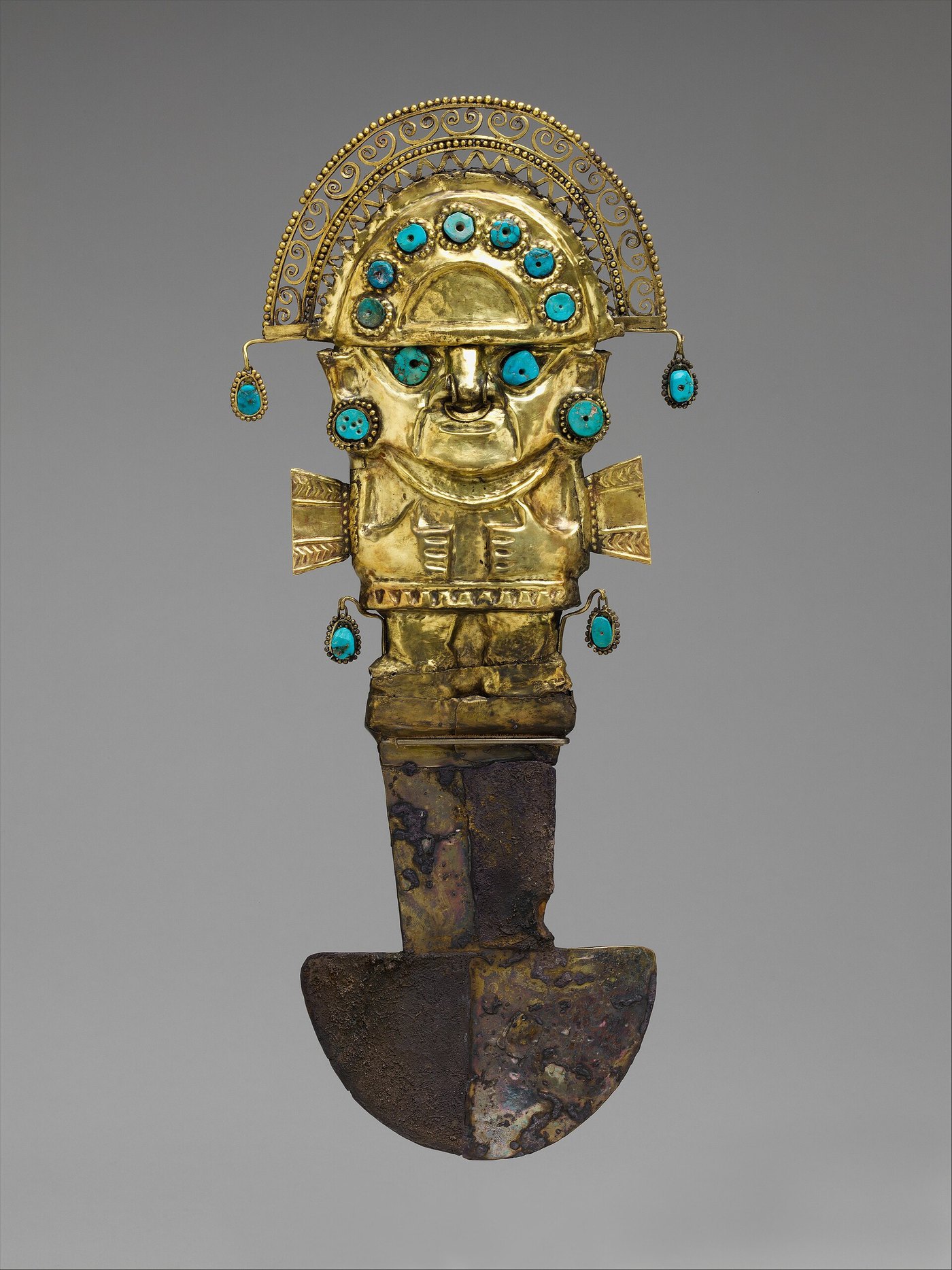 An ornate ceremonial knife against a grey background: the upper handle is a gold figure of a deity with an arched, filigreed headdress and turquoise inlays, above a tarnished crescent-shaped metal blade