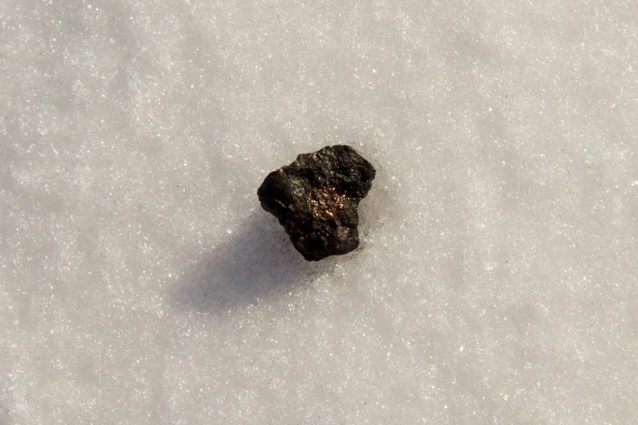 A single small dark angular stone with a blackened crust lying on bright sunlit snow, casting a short shadow, with nothing else in the frame