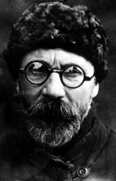 A black and white portrait of a bearded man in round wire-rimmed spectacles and a fur hat, wearing a heavy coat with a large button, looking directly at the camera