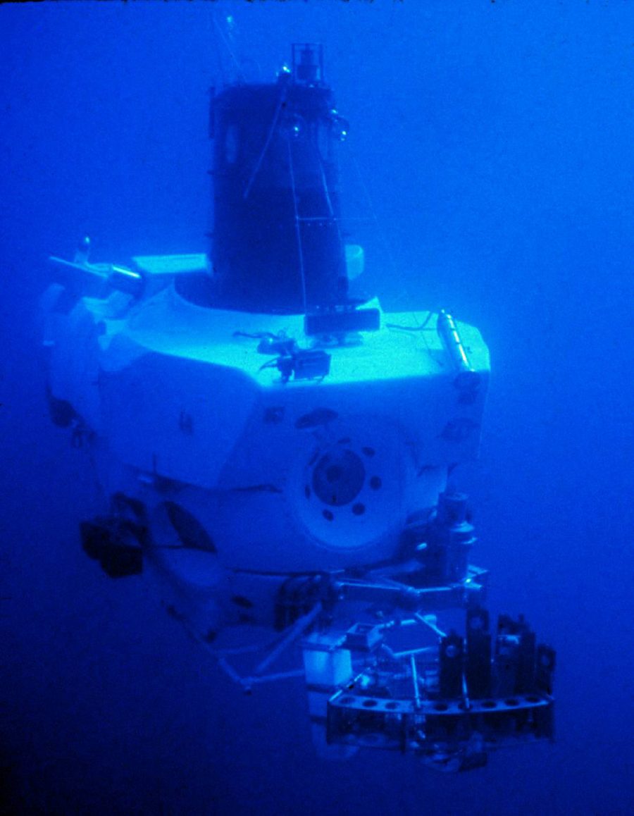 The submersible Alvin descending through blue water, seen from above and behind. A white pressure-hull body with a dark conning tower, an external instrument frame, and sample baskets and manipulator gear mounted on the front, in strong blue-filtered light.
