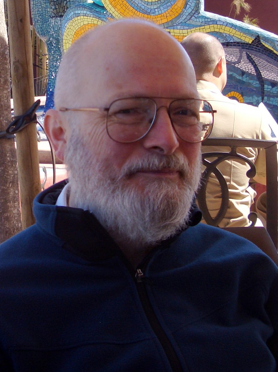A bald, white-bearded man in wire-framed glasses and a dark navy fleece jacket, seated outdoors in sunlight in front of a coloured mosaic wall: Vernor Vinge, photographed in March 2008