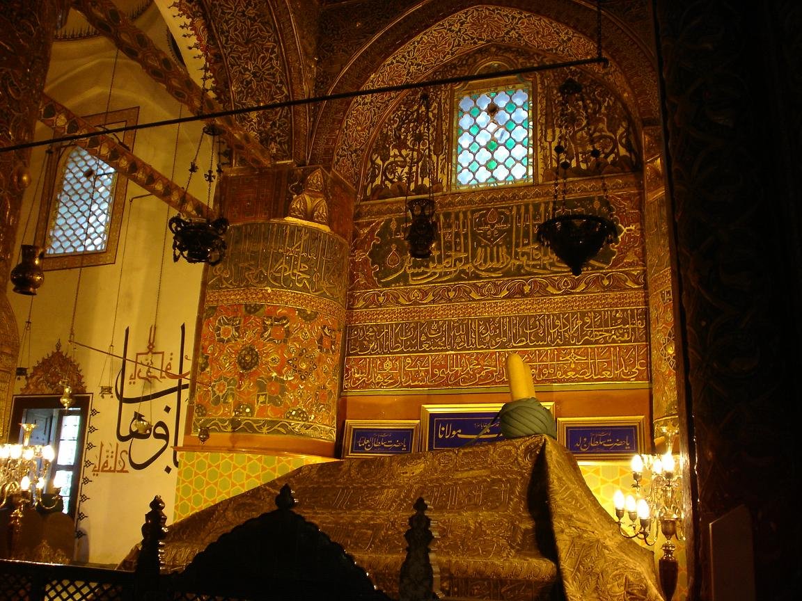 The ornate interior of Rumi's tomb chamber at the Mevlana Museum in Konya, with gold calligraphy covering the walls, hanging lamps, stained-glass windows, and a draped sarcophagus topped by a large green turban