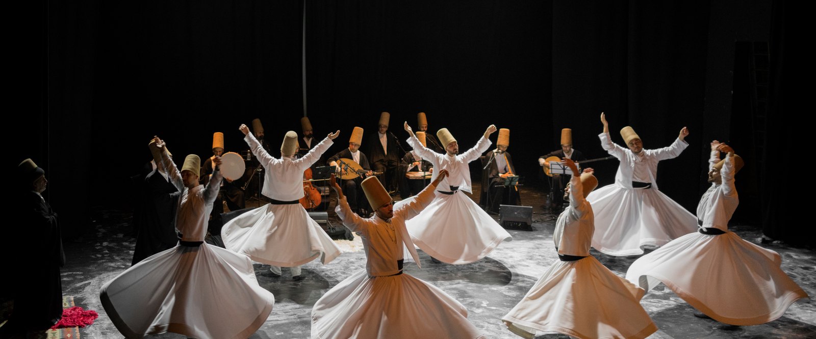 Whirling dervishes of the Mevlevi Order in white flared skirts and tall camel-hair hats performing the Sema, caught mid-spin before seated musicians