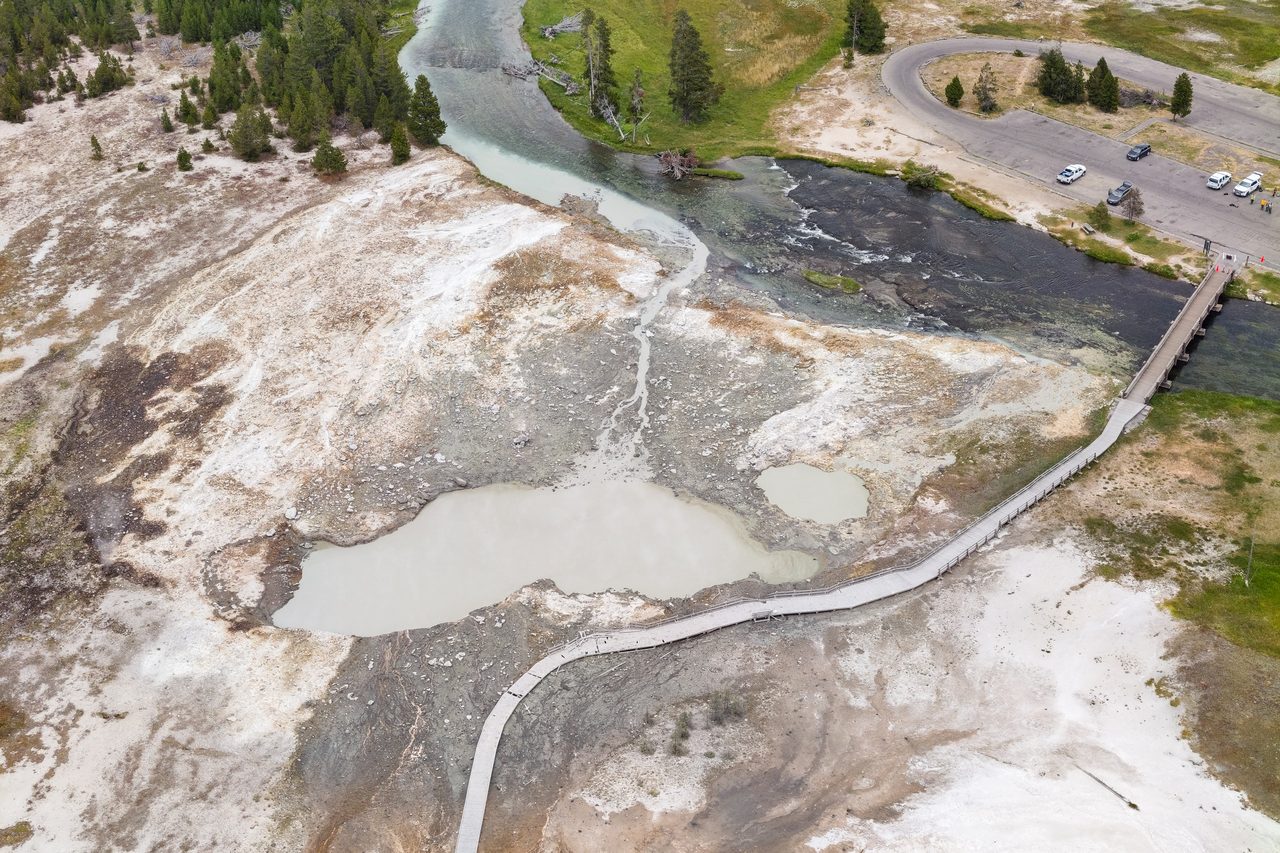 An aerial photograph of the Biscuit Basin debris field: a grey blast deposit spread across pale sinter, a broken boardwalk running through it, a milky crater pool, and a river and a parking lot beyond