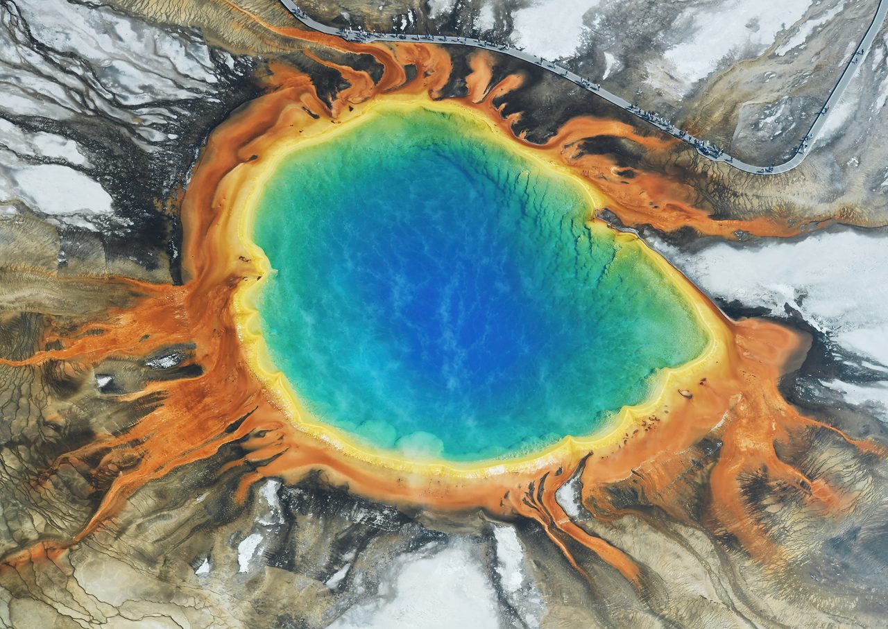 An aerial view looking straight down onto Grand Prismatic Spring, a deep blue pool ringed by yellow, orange and rust-colored microbial mats spreading outward across pale sinter